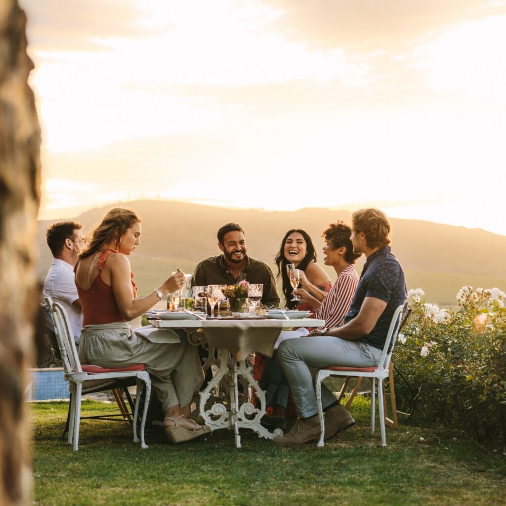 Group of friends enjoying an outdoor sunset dinner with drinks in a scenic countryside setting”