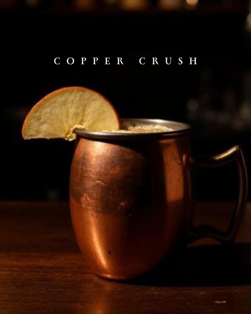 Copper mug cocktail called Copper Crush garnished with a dried citrus slice on a wooden bar.